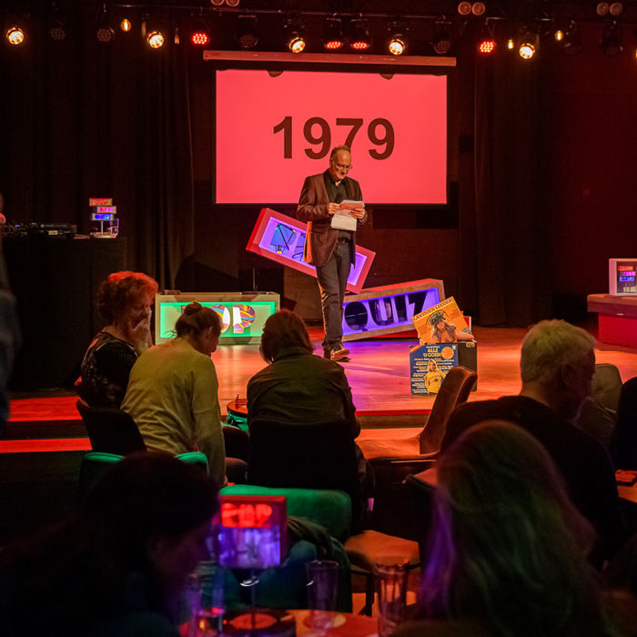 Jeroen (presentator Pop Bizz Quiz) op het podium met microfoon in hand en deel van het publiek