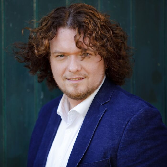 Zanger Jason Bouman staart in de camera. Hij heeft bruine krullen en draagt een blauw jasje en wit overhemd.