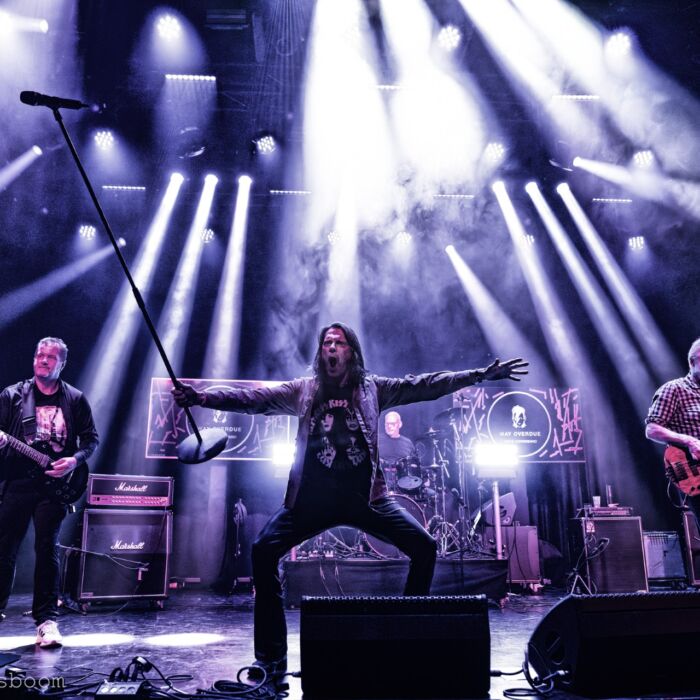 Zanger van Way Overdue poseert met gebogen benen en gespreide armen op het podium met microfoonstandaard in de hand.