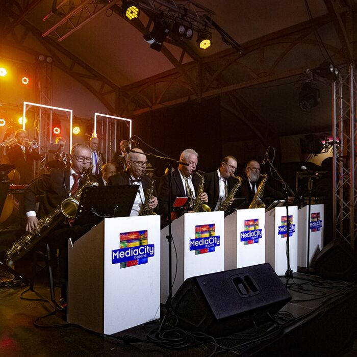 Mediacity BigBand performen zittend achter witte decorkastjes op hun blaasinstrumenten.