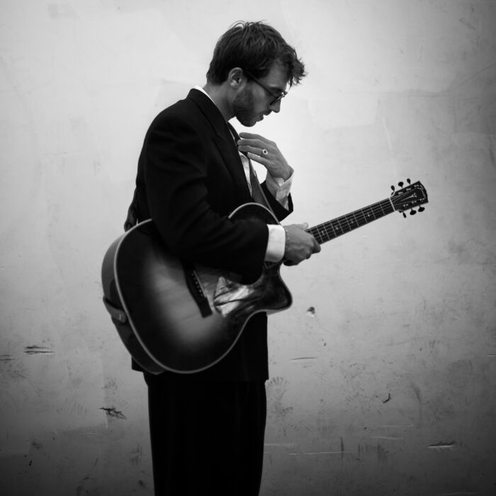 Zwart wit foto van Ground Up artiest Romeo Marino, met een zonnebril op en een gitaar in zijn hand.