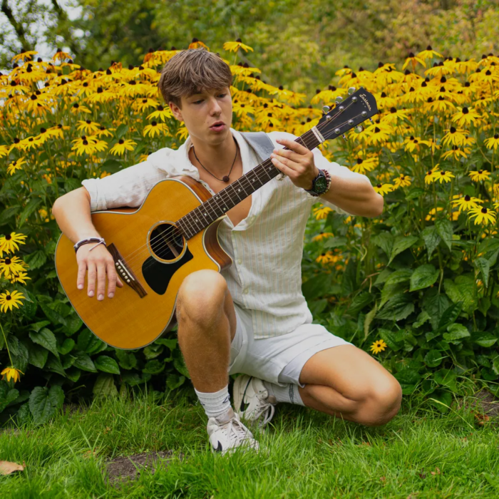 Ground Up artiest Victor Germanus knielt in het gras met zonnebloemen op de achtergrond en een gitaar in zijn hand.
