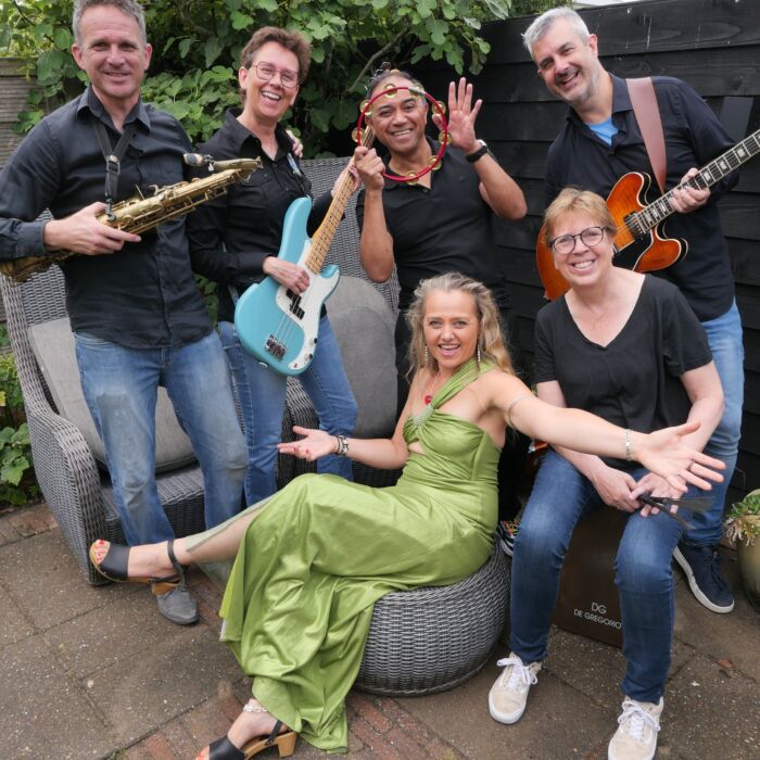 Bandleden van The Living Room Project poseren lachend en vrolijk voor de camera in een achtertuin, met instrumenten in de hand.
