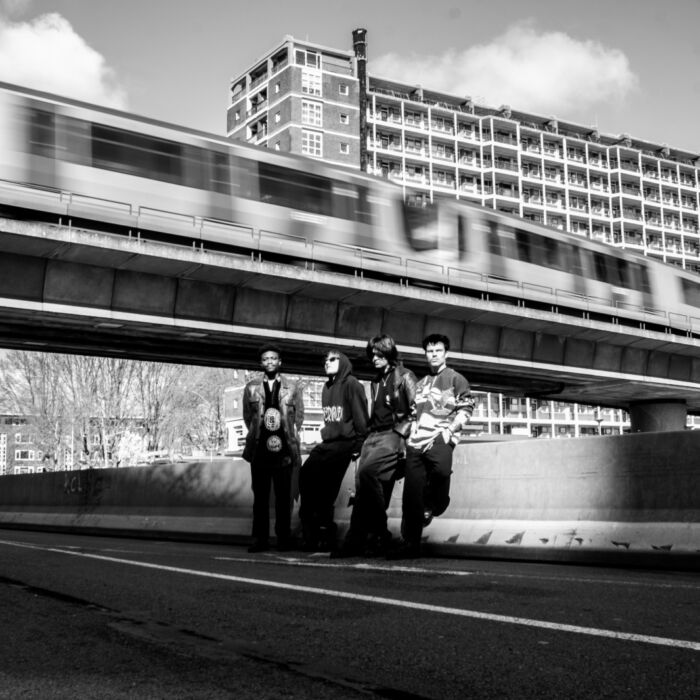 Zwartwitfoto van bandleden Loveth Besamoh. Ze staan op een industriële plek voor een voorbijrijdende trein.