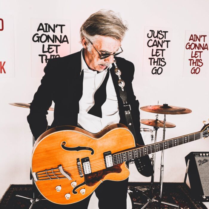 Muzikant Glen Matlock staart naar beneden met zonnebril op en houten semi hollow gitaar om zijn nek en handen in zijn zakken.
