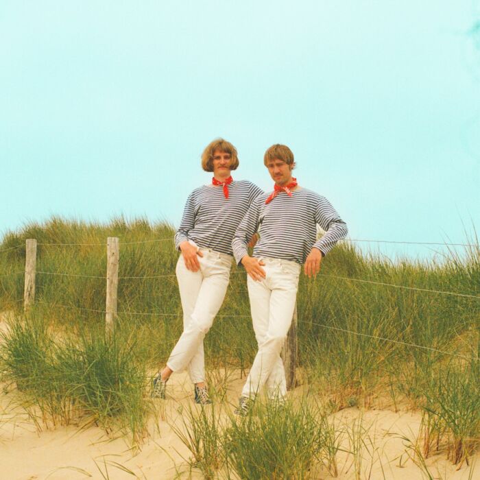 op de foto zijn de 2 artiesten te zien die in de duinen staan leunden tegen een hekje met een blauwe lucht als achtergrond
