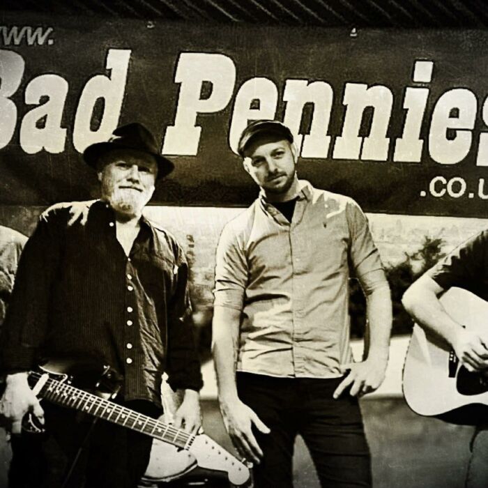 Zwart-witfoto van bluesrockband Bad Pennies uit Engeland. Ze poseren met instrument in de hand voor een banner met hun bandnaam erop.