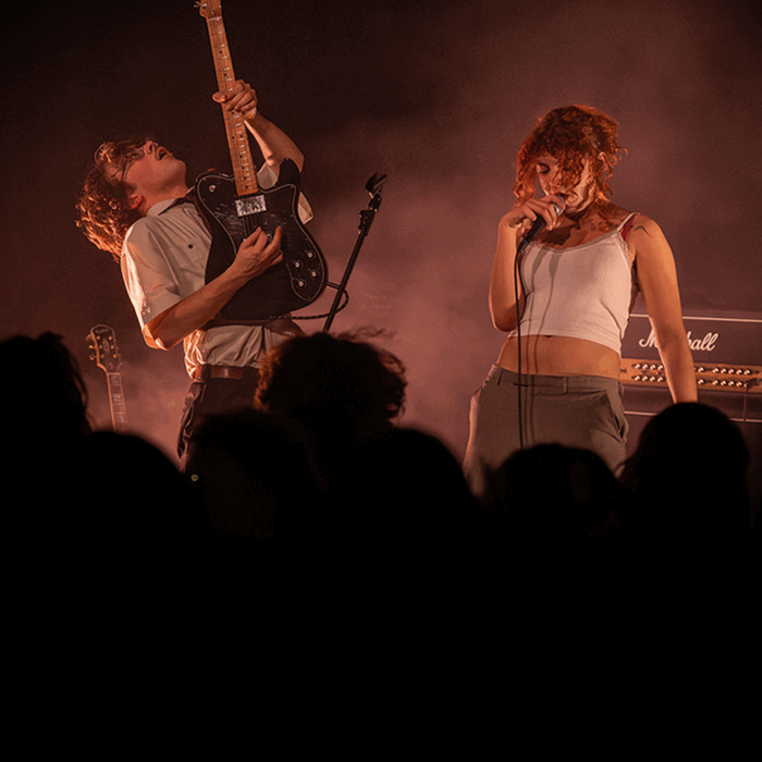 Bandleden van punkband clitteband performen expressief voor volle zaal. Zangeres en gitarist zijn in beeld.