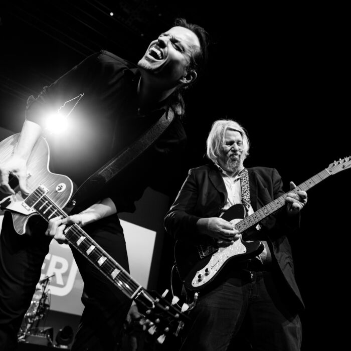 De foto is zwart wit. Op de foto zijn 2 band leden van Cooking With Knopfler te zien, ze spelen alle 2 met een gitaar en staan te zingen.