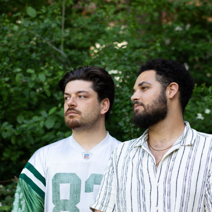 Rapduo Bandietos kijkt synchroon dezelfde zijrichting op. Op de achtergrond staat een groene struik.