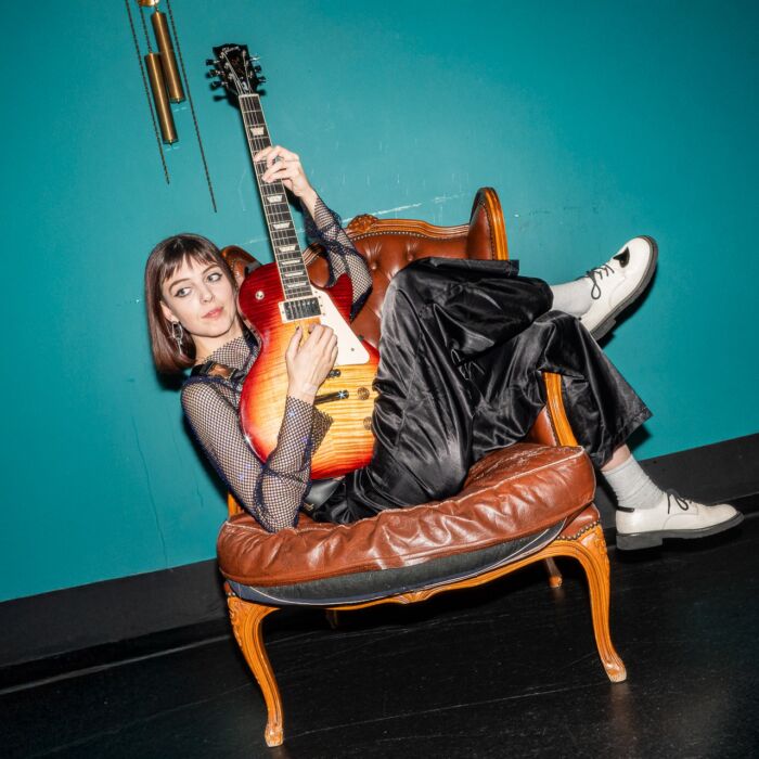 Artiest Inge Lamboo hangt over bruine leren stoel met benen over linker armleuning en sunburst-kleurige elektrische gitaar in de hand.