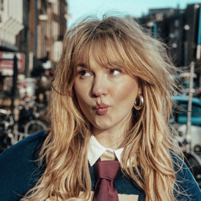 Artiest Judith tuit haar lippen en kijkt speels omhoog. Ze draagt een stropdas over haar trui.