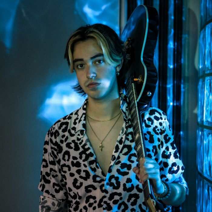 Artiest Zikko Lopez staart in de camera met panterprintbloes aan, zijn gitaar hangt over zijn linkerschouder.