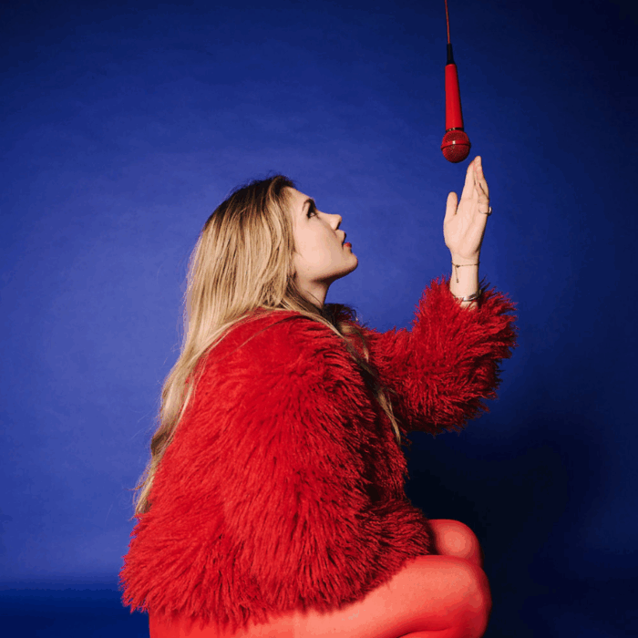 Zangeres minke poseert in rode outfit voor blauwe achtergrond. Gehurkt op de grond kijkt ze naar een rode microfoon die voor haar neus hangt.