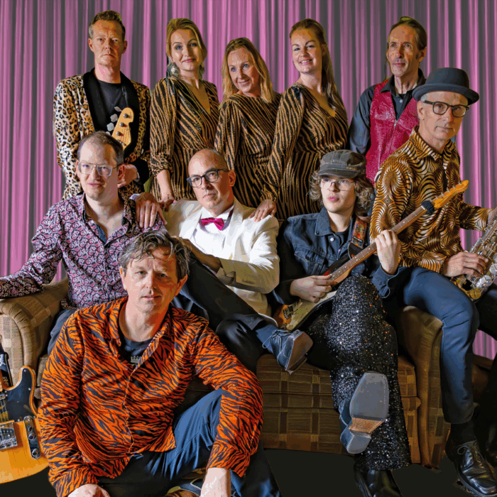 De tien bandleden van More Than This zitten op en voor een bank, sommigen staan achter de bank. Iedereen kijkt vrolijk in de camera.