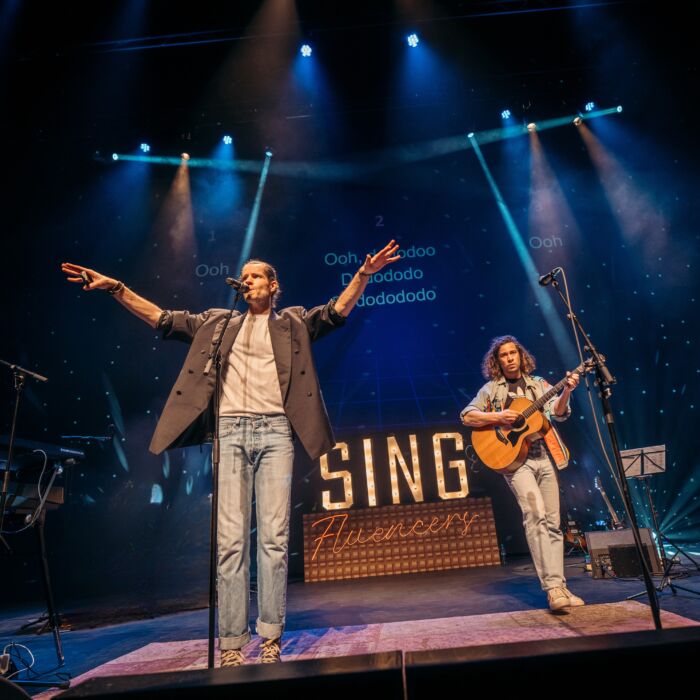 op de foto zijn de 2 artiesten van Popup Choir te zien op een podium. eentje staat met een gitaar en de andere staat te zingen.