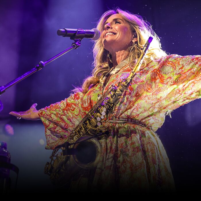 Saxofoniste Candy Dulfer staat met open armen lachend op het podium, haar saxofoon hangt om haar nek.