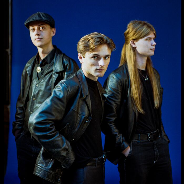 Drie bandleden van Helen Jewett poseren voor donkerblauwe achtergrond in zwarte leren kleding.