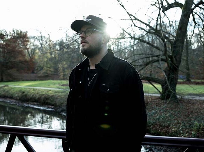 Artiest Prize Collect staart in de verte poserend in een park. Hij draagt donkere kleding en een pet en een bril.