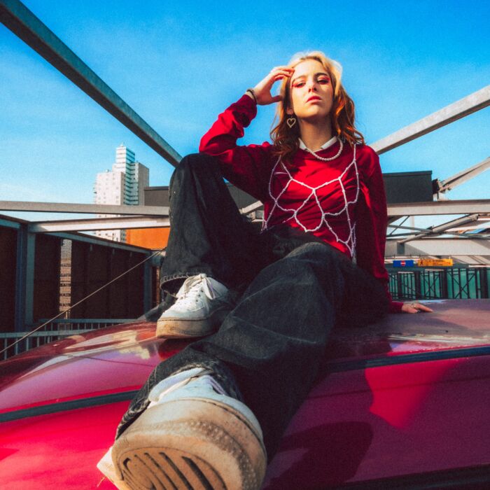 Artiest XXJULIA zit achterover op een rode auto en leunt op linkerarm.