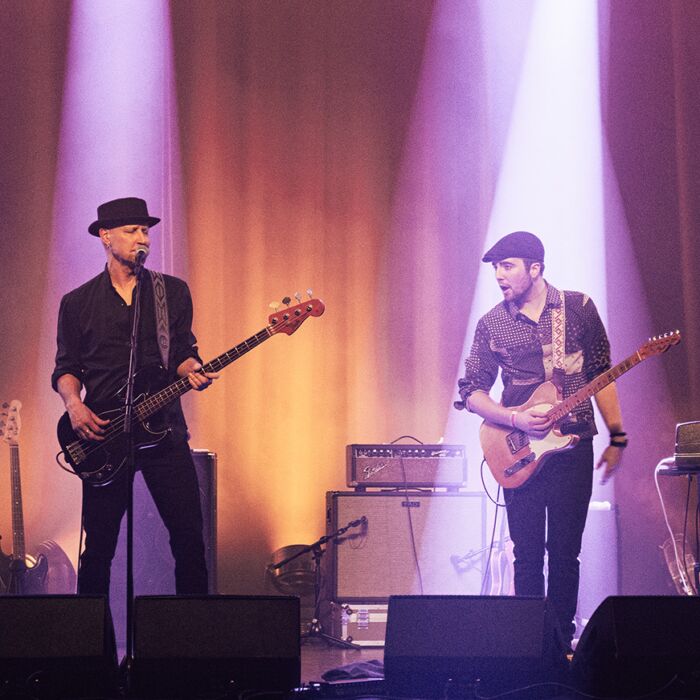 Vier muzikanten spelen live op een podium met felle oranje en paarse spotlights. Je ziet een drummer, bassist, gitarist en toetsenist, omringd door rook en warme podiumverlichting.
