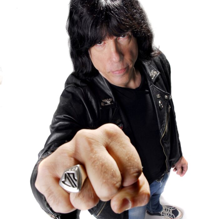Ramones-drummer Marky Ramone poseert voor de camera in witte studio met vuist richting de camera. Hij draagt een zwarte leren jas en een spijkerbroek met gympen.