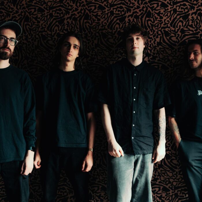 Bandleden van Sugar Spine poseren in zwarte kleding tegen zwarte achtergrond. Alleen hun hoofden en armen vallen op in het licht.