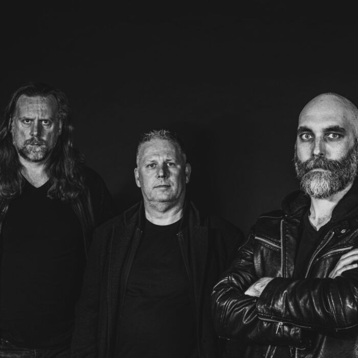 De drie bandleden van Chaos Unleashed in het zwart gekleed tegen een zwarte achtergrond