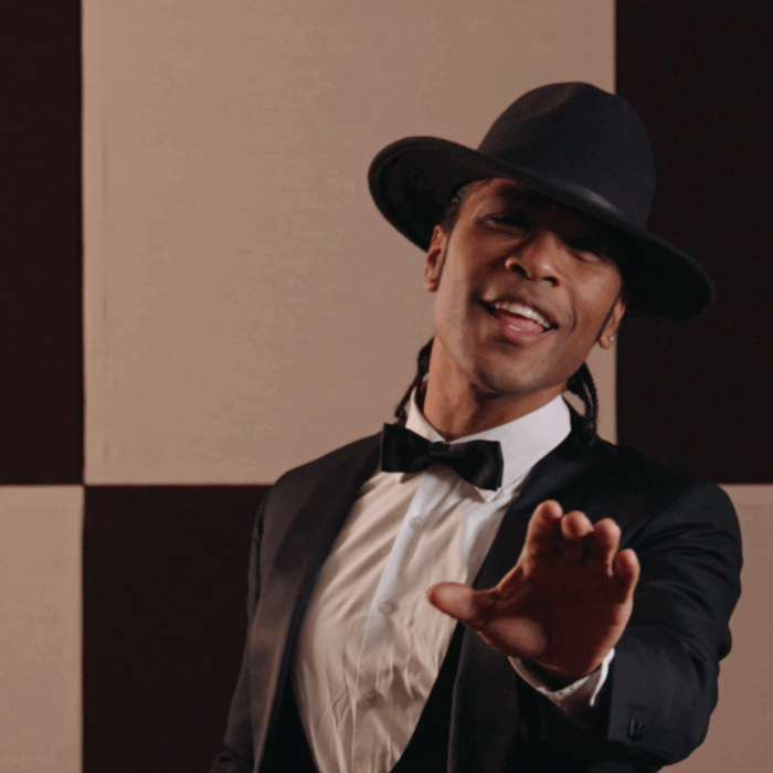 Jared Grant heeft een pak aan met een hoed op en staat te zingen voor de camera