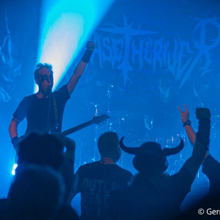 Metalcafe sfeerimpressie foto. Op de afbeelding zie je donkere silhouetten van muzikanten op het podium en juichend publiek.
