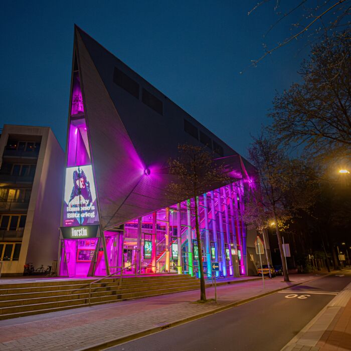 De buitenkant van De Vorstin in het avondlicht met de verlichting van het pand in alle kleuren van de regenboog