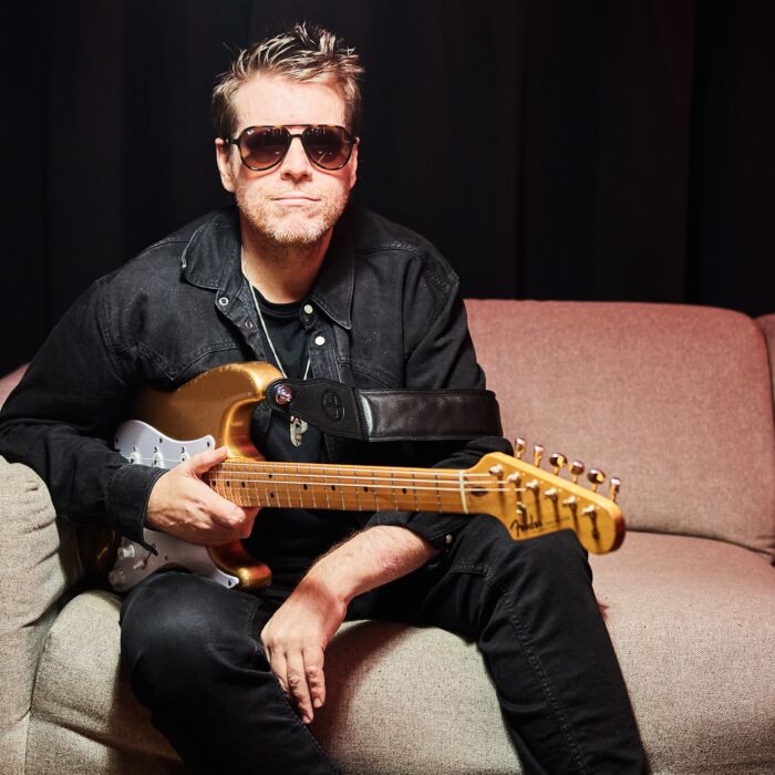 Danny Bryant zit met zijn houtkleurige Fender Stratocaster op een witte bank